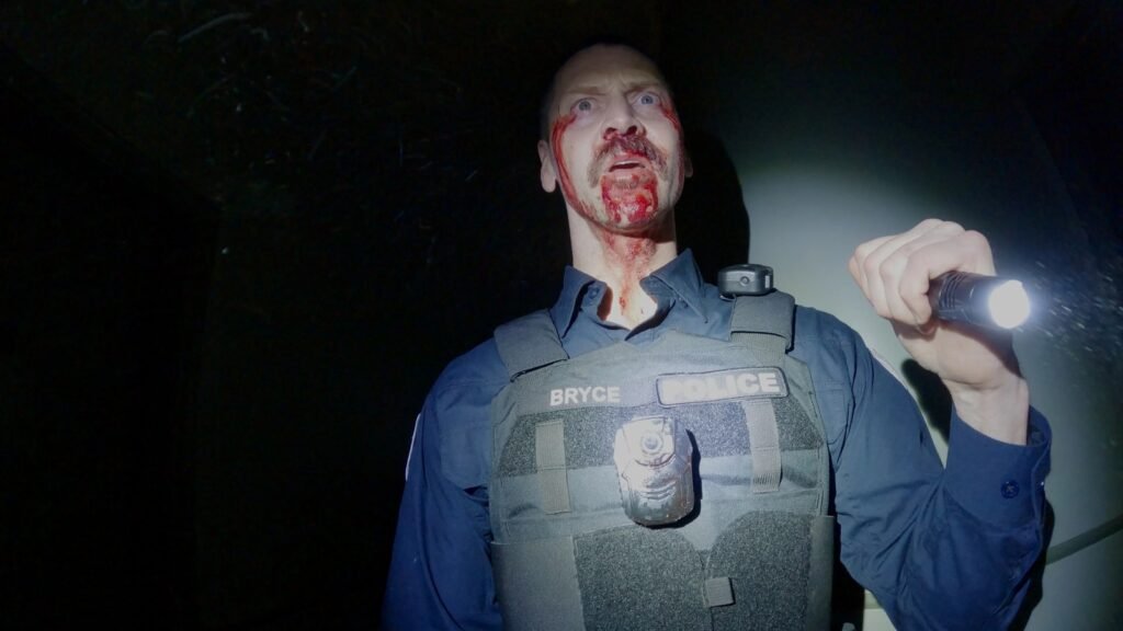 Sean Rogerson as Officer Bryce Anderson, bloodied and holding a flashlight, his chest-mounted body camera lit, in Bodycam (2026)