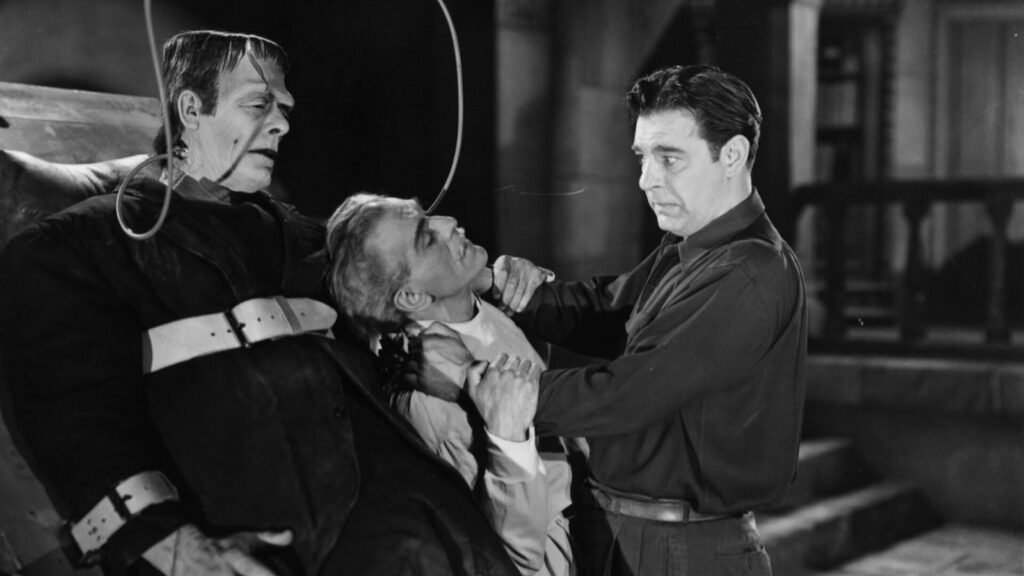 Frankenstein's Monster (Glenn Strange) strapped to the laboratory table gripping Dr. Gustav Niemann (Boris Karloff), as Lawrence Talbot (Lon Chaney Jr.) pulls him free, in House of Frankenstein (1944)
