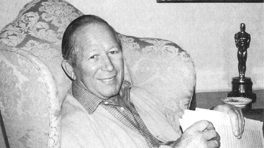 Black-and-white portrait of composer James Bernard seated in an armchair holding manuscript pages, his Academy Award statuette on the table beside him