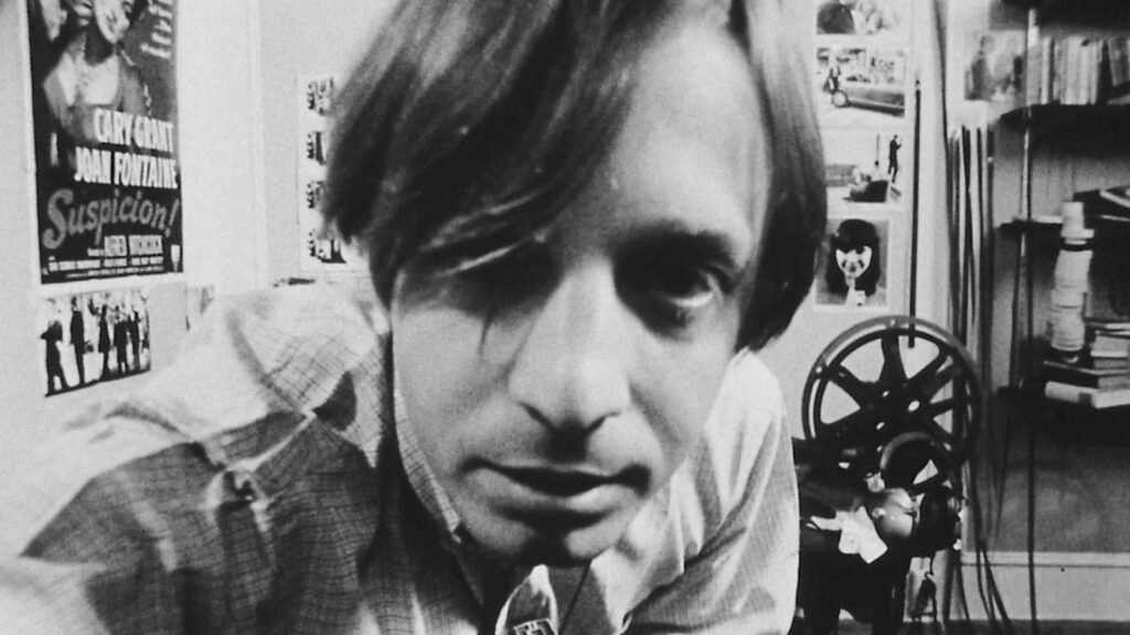L. M. Kit Carson as David Holzman, leaning into his own camera in a black-and-white still from David Holzman's Diary (1967), a Suspicion poster and film projector behind him