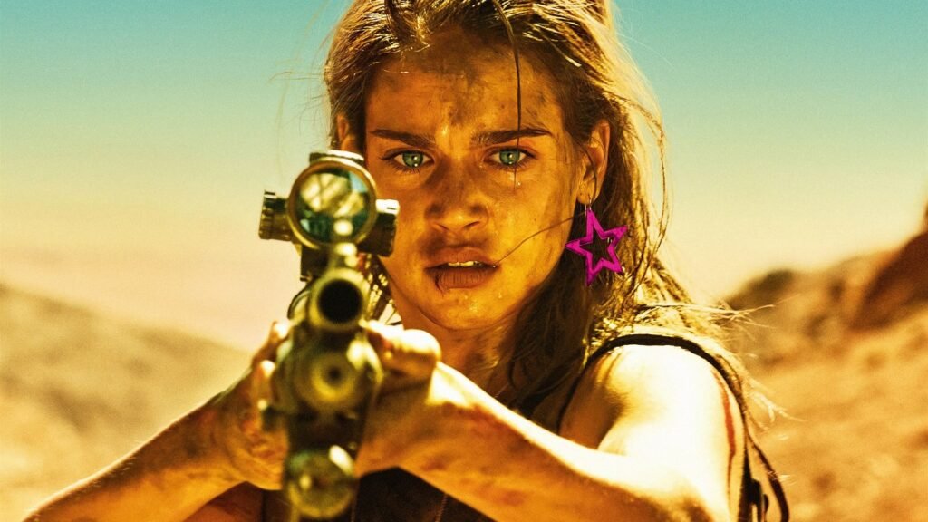 Matilda Lutz as Jen in Revenge (2017) — dirt- and blood-streaked, aiming a scoped rifle straight at camera in the desert, wearing the character's pink star earring.