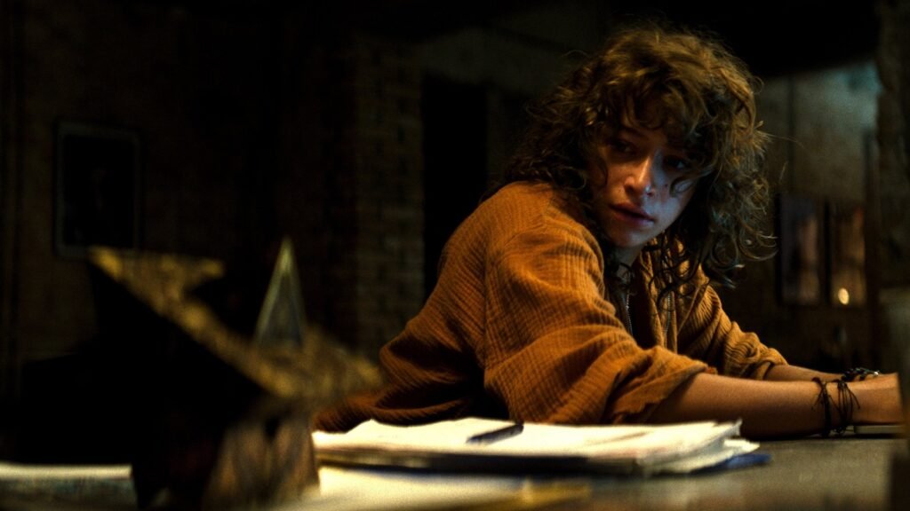 Odessa A’zion as Riley McKendry in Hellraiser (2022), seated at a table with the puzzle box in the foreground