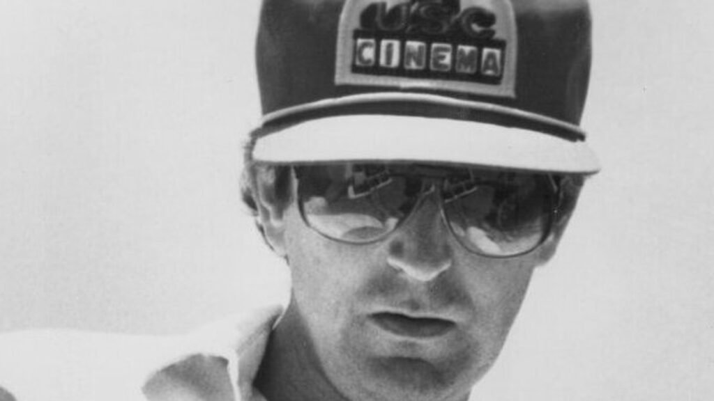 Black-and-white portrait of director Richard Franklin wearing sunglasses and a USC Cinema cap, holding a copy of Psycho