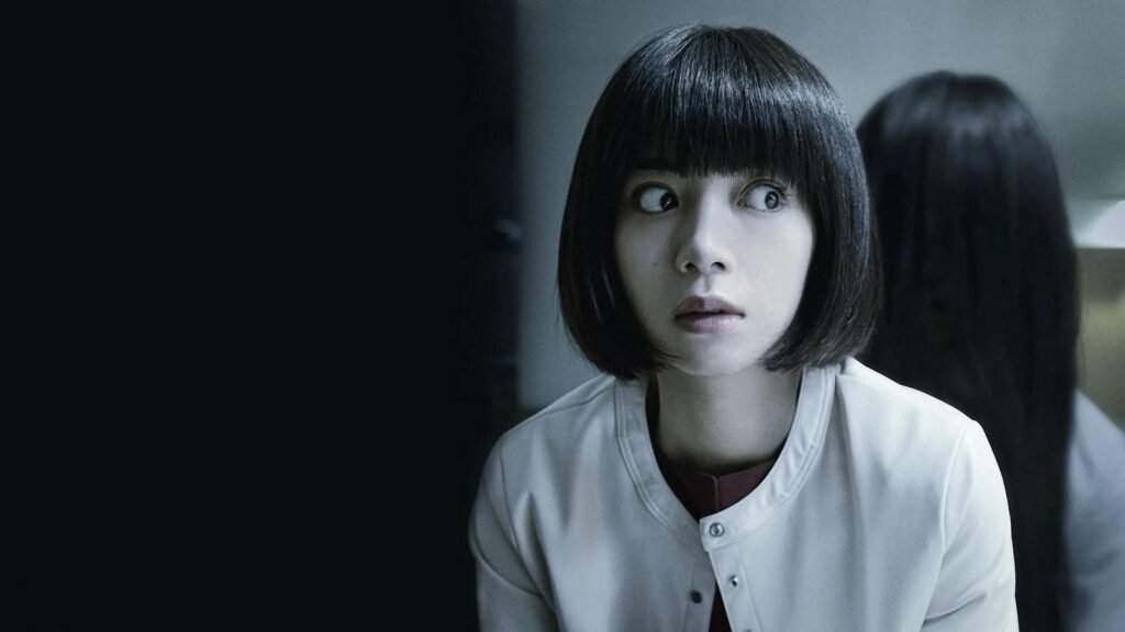 Elaiza Ikeda as Mayu Akikawa, staring in fright as Sadako's long-haired figure stands behind her, in Sadako (2019)