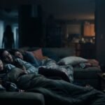 Three friends asleep on the couch at a sleepover while a shadowy figure looms in the dark hallway — Blumhouse's Sleepover