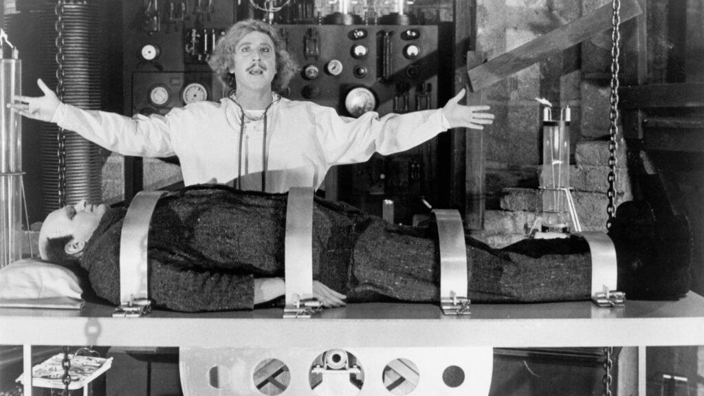 Gene Wilder as Frederick Frankenstein, arms spread over Peter Boyle's Monster strapped to the laboratory table in Young Frankenstein (1974)